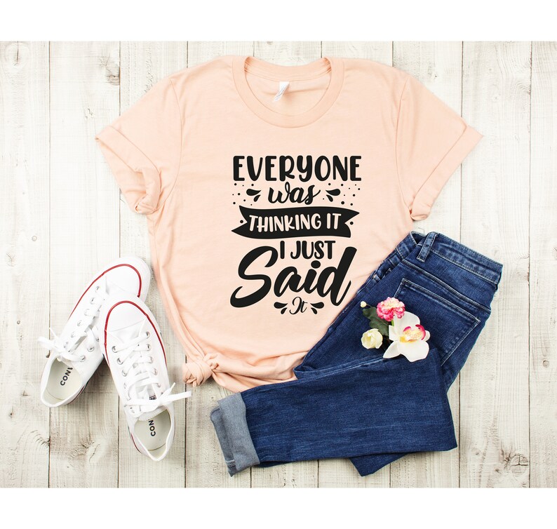May include: A peach-colored t-shirt with the text "Everyone was thinking it I just said it" in black lettering. The shirt is paired with blue jeans and white sneakers.
