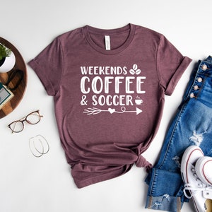 Op de afbeelding: Een maronkleurig T-shirt met de tekst "Weekends Coffee & Soccer" in wit, met een koffiekopje-icoon en een pijl met een hart.