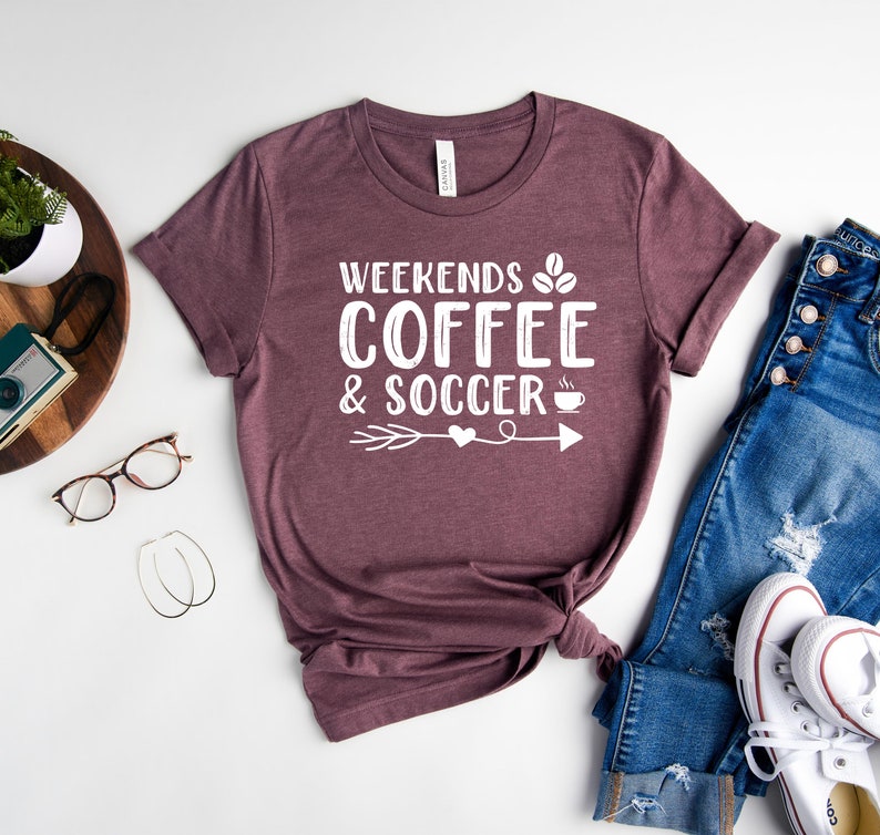 Op de afbeelding: Een maronkleurig T-shirt met de tekst "Weekends Coffee & Soccer" in wit, met een koffiekopje-icoon en een pijl met een hart.