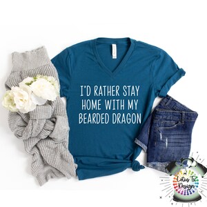 Puede incluir: Camiseta azul turquesa de cuello en V con el texto "I'd rather stay home with my bearded dragon" en blanco. La camiseta est&aacute; colocada sobre una superficie blanca con un su&eacute;ter gris y unos pantalones cortos de mezclilla azul.