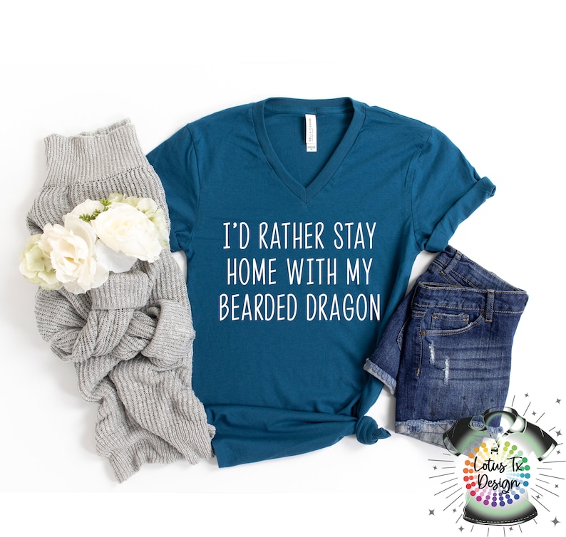 Puede incluir: Camiseta azul turquesa de cuello en V con el texto "I'd rather stay home with my bearded dragon" en blanco. La camiseta est&aacute; colocada sobre una superficie blanca con un su&eacute;ter gris y unos pantalones cortos de mezclilla azul.