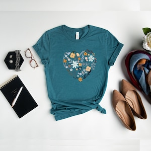 K&ouml;nnte beinhalten: Tealblaues T-Shirt mit einem floralen Herzdesign. Das Herz besteht aus verschiedenen Blumen in Blaut&ouml;nen, Gelb und Orange.