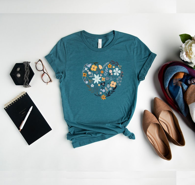 K&ouml;nnte beinhalten: Tealblaues T-Shirt mit einem floralen Herzdesign. Das Herz besteht aus verschiedenen Blumen in Blaut&ouml;nen, Gelb und Orange.