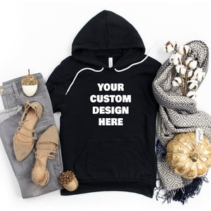 Może przedstawiać: Czarna bluza z kapturem z białą kieszenią i nadrukiem "YOUR CUSTOM DESIGN HERE" z przodu. Bluza leży płasko na białym tle z parą brązowych sandał&oacute;w, szarymi dżinsami, ozdobnym żołędziem, białą gałązką bawełny z szalem w szaro-białe wzory i złotą dynią.