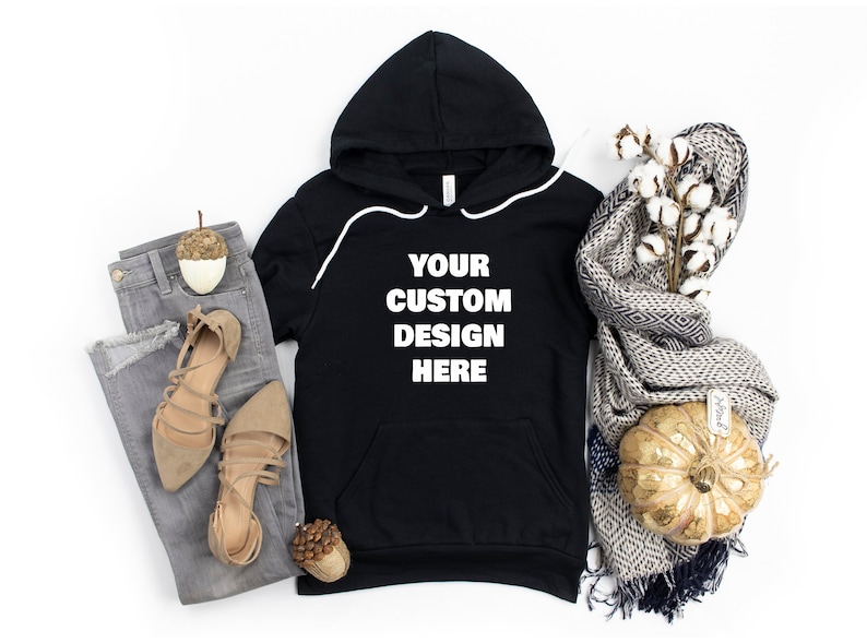 Może przedstawiać: Czarna bluza z kapturem z białą kieszenią i nadrukiem "YOUR CUSTOM DESIGN HERE" z przodu. Bluza leży płasko na białym tle z parą brązowych sandał&oacute;w, szarymi dżinsami, ozdobnym żołędziem, białą gałązką bawełny z szalem w szaro-białe wzory i złotą dynią.