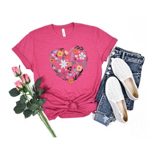 K&ouml;nnte beinhalten: Ein pinkfarbenes T-Shirt mit einem floralen Herzdesign. Das Herz besteht aus bunten Blumen und Bl&auml;ttern. Das Shirt wird mit einer blauen Jeans und wei&szlig;en Schuhen kombiniert.