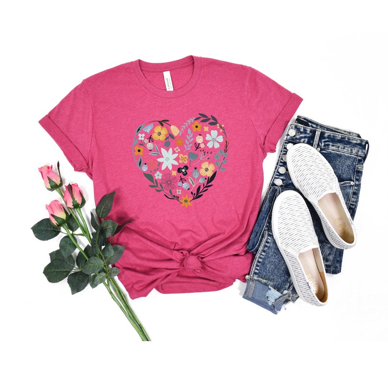 K&ouml;nnte beinhalten: Ein pinkfarbenes T-Shirt mit einem floralen Herzdesign. Das Herz besteht aus bunten Blumen und Bl&auml;ttern. Das Shirt wird mit einer blauen Jeans und wei&szlig;en Schuhen kombiniert.