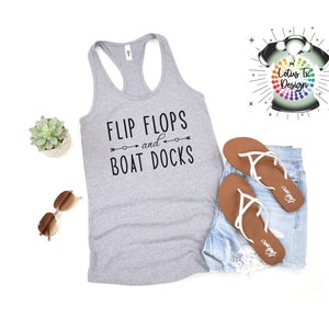 May include: Gray tank top with the text "Flip Flops and Boat Docks" in black. The tank top is laying on a white surface with a pair of brown flip flops and denim shorts.