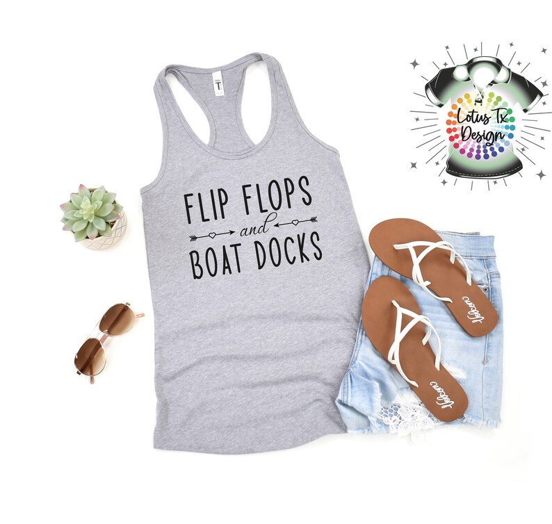 May include: Gray tank top with the text "Flip Flops and Boat Docks" in black. The tank top is laying on a white surface with a pair of brown flip flops and denim shorts.