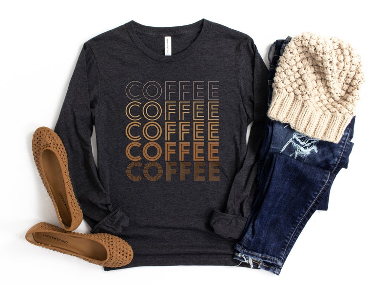 K&ouml;nnte beinhalten: Ein dunkelgraues Langarmshirt mit dem Wort "COFFEE" f&uuml;nfmal wiederholt in einem Farbverlauf von Braunt&ouml;nen. Das Shirt wird mit einer blauen Jeans, einer cremefarbenen M&uuml;tze und braunen Ballerinas gestylt.