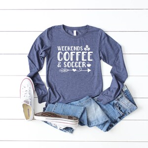 Op de afbeelding: Een marineblauw longsleeve shirt met de tekst "Weekends Coffee & Soccer" in wit, met een afbeelding van een koffiekopje en een voetbal. Het shirt ligt op een wit houten oppervlak met een paar witte sneakers en een blauwe jeans.