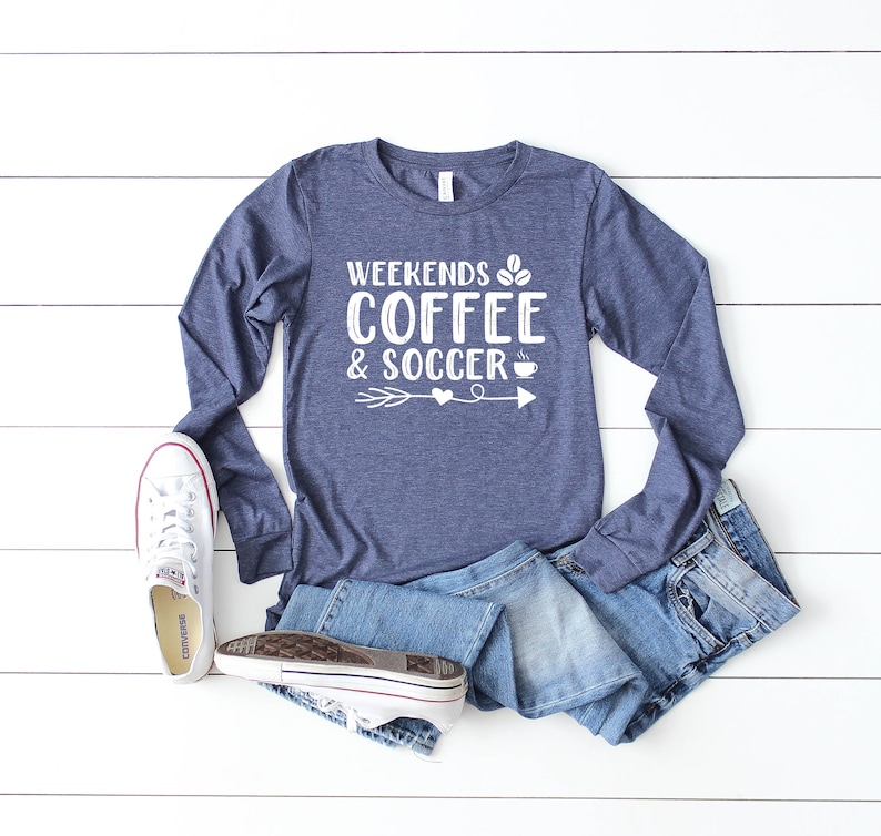 Op de afbeelding: Een marineblauw longsleeve shirt met de tekst "Weekends Coffee & Soccer" in wit, met een afbeelding van een koffiekopje en een voetbal. Het shirt ligt op een wit houten oppervlak met een paar witte sneakers en een blauwe jeans.