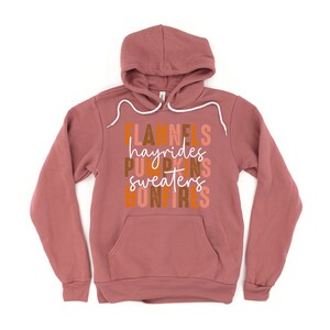 May include: A mauve hoodie with a drawstring hood and a front pocket. The front of the hoodie features the words "Flannels, hayrides, pumpkins, sweaters, bonfires" in a stacked, colourful design. The hoodie is made of a soft material.