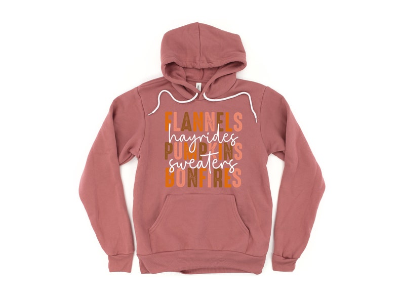 May include: A mauve hoodie with a drawstring hood and a front pocket. The front of the hoodie features the words "Flannels, hayrides, pumpkins, sweaters, bonfires" in a stacked, colourful design. The hoodie is made of a soft material.