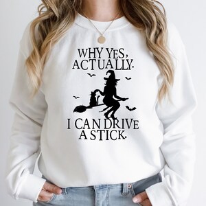 May include: White sweatshirt with a black graphic of a witch flying on a broomstick with a cat. The text reads "Why yes, actually. I can drive a stick."