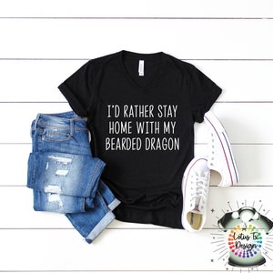 Puede incluir: Camiseta negra de cuello en V con el texto "I'd rather stay home with my bearded dragon" impreso en blanco en la parte delantera. La camiseta se combina con jeans azules y zapatillas blancas.