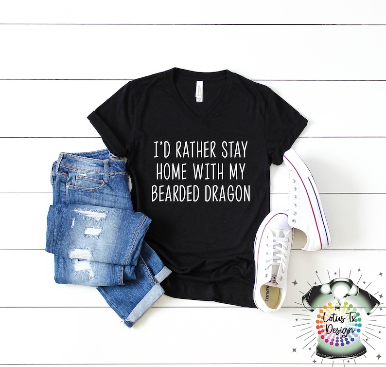 Puede incluir: Camiseta negra de cuello en V con el texto "I'd rather stay home with my bearded dragon" impreso en blanco en la parte delantera. La camiseta se combina con jeans azules y zapatillas blancas.