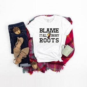 May include: White t-shirt with the text "Blame it all on my roots" in black lettering. A small cartoon boot with a cowboy hat is above the word "roots". The shirt is laid out on a red and white plaid blanket with a pair of brown sandals, a pair of blue jeans, and a green mug.