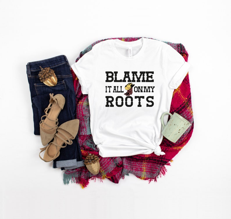 May include: White t-shirt with the text "Blame it all on my roots" in black lettering. A small cartoon boot with a cowboy hat is above the word "roots". The shirt is laid out on a red and white plaid blanket with a pair of brown sandals, a pair of blue jeans, and a green mug.