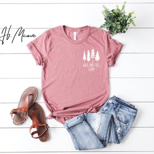May include: A mauve colored t-shirt with a white graphic of trees and the text "Wild and Free Camp". The t-shirt is laid out on a white wooden surface with a pair of brown sandals and blue jeans.