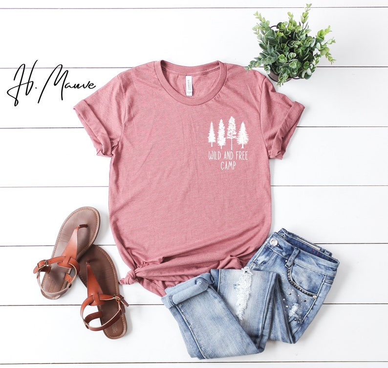 May include: A mauve colored t-shirt with a white graphic of trees and the text "Wild and Free Camp". The t-shirt is laid out on a white wooden surface with a pair of brown sandals and blue jeans.