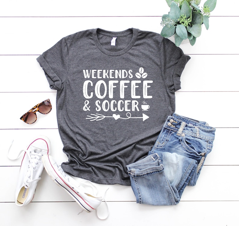 Op de afbeelding: Een donkergrijs T-shirt met de tekst "Weekends Coffee & Soccer" in wit, met een koffiekopje, koffiebonen en een pijl met een hart.