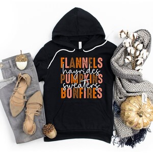 May include: Black hooded sweatshirt with the words "FLANNELS hayrides PUMPKINS sweaters BONFIRES" in orange, brown, and white. The sweatshirt is paired with grey jeans, tan sandals, and a decorative scarf.