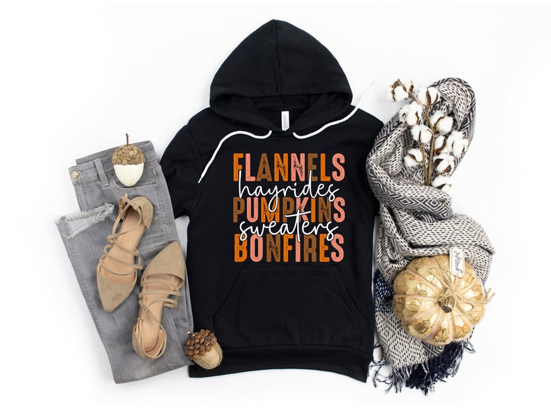May include: Black hooded sweatshirt with the words "FLANNELS hayrides PUMPKINS sweaters BONFIRES" in orange, brown, and white. The sweatshirt is paired with grey jeans, tan sandals, and a decorative scarf.