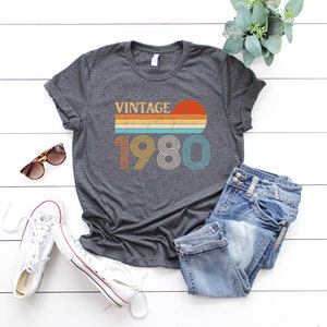 May include: A dark gray t-shirt with a vintage 1980 design. The design features a sunset with a rainbow stripe and the year 1980 in a retro font.