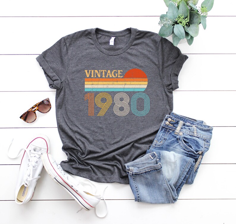 May include: A dark gray t-shirt with a vintage 1980 design. The design features a sunset with a rainbow stripe and the year 1980 in a retro font.