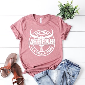 May include: A dusty rose t-shirt with a white graphic of a bull skull with the text "Try That In A Small Town" and the word "ALDEAN" in a stylized font.