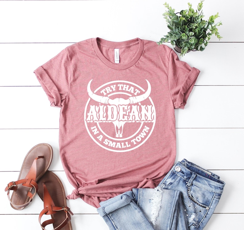 May include: A dusty rose t-shirt with a white graphic of a bull skull with the text "Try That In A Small Town" and the word "ALDEAN" in a stylized font.