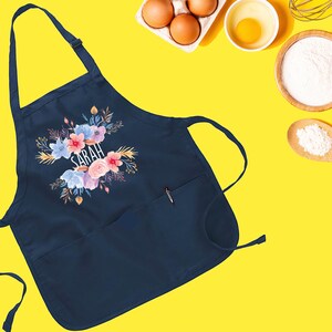May include: A navy blue apron with a floral design and the name "SARAH" printed in white. The apron has a large pocket on the front.