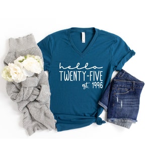 May include: Teal blue v-neck t-shirt with the text "hello twenty-five est. 1996" in white. The shirt is laid flat on a white background with a gray sweater and denim shorts.