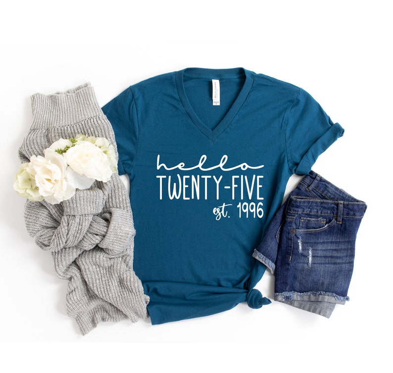 May include: Teal blue v-neck t-shirt with the text "hello twenty-five est. 1996" in white. The shirt is laid flat on a white background with a gray sweater and denim shorts.