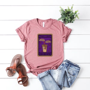 Puede incluir: Una camiseta rosa con un gr&aacute;fico de una carta de tarot que presenta una taza de caf&eacute; helado con el texto "The Iced Coffee".