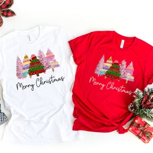 Puede incluir: Dos camisetas con un dise&ntilde;o de &aacute;rbol de Navidad. La camiseta blanca tiene un dise&ntilde;o de &aacute;rbol rosa, verde y dorado con el texto "Merry Christmas". La camiseta roja tiene el mismo dise&ntilde;o con el texto "Merry Christmas" en blanco.