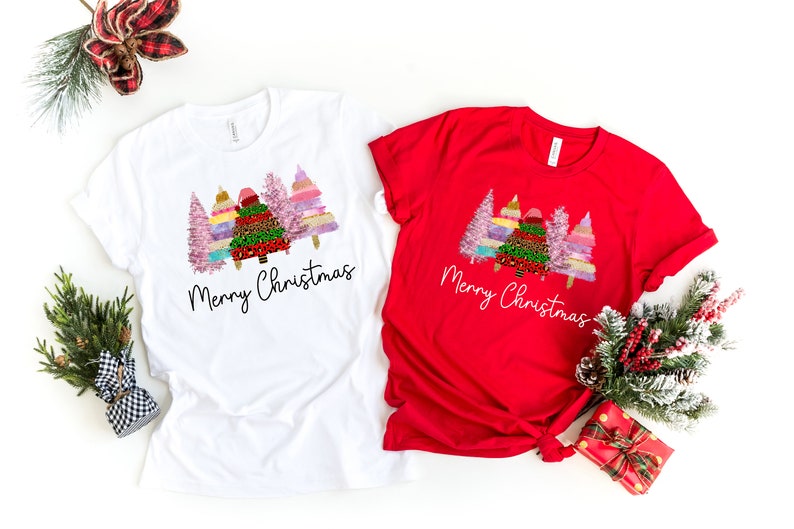 Puede incluir: Dos camisetas con un dise&ntilde;o de &aacute;rbol de Navidad. La camiseta blanca tiene un dise&ntilde;o de &aacute;rbol rosa, verde y dorado con el texto "Merry Christmas". La camiseta roja tiene el mismo dise&ntilde;o con el texto "Merry Christmas" en blanco.