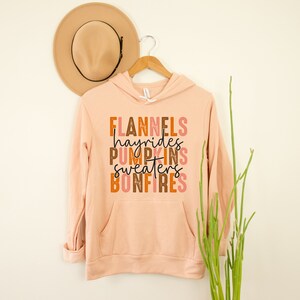 May include: A peach-coloured hoodie with a front pocket and the words "FLANNELS hayrides PUMPKINS SWEATERS BONFIRES" in a fall-themed colour palette. A tan hat hangs above the hoodie on a wooden hanger.