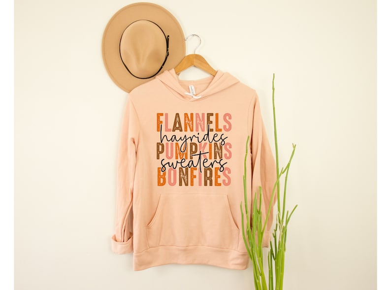 May include: A peach-coloured hoodie with a front pocket and the words "FLANNELS hayrides PUMPKINS SWEATERS BONFIRES" in a fall-themed colour palette. A tan hat hangs above the hoodie on a wooden hanger.