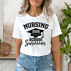 May include: White t-shirt with the text "NURSING 2026 school Survivor" in black. The design includes a graduation cap. The model is wearing blue denim jeans.
