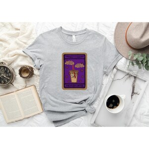 Puede incluir: Una camiseta gris con un dise&ntilde;o de carta de tarot morado y marr&oacute;n que presenta un caf&eacute; helado de dibujos animados con una pajita roja y el texto "The Iced Coffee".