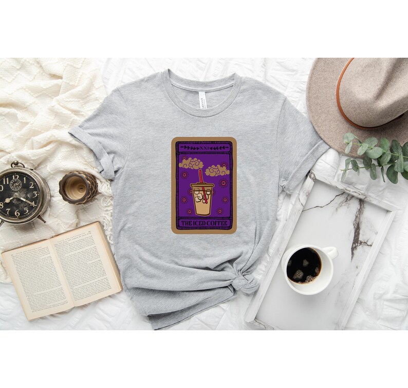 Puede incluir: Una camiseta gris con un dise&ntilde;o de carta de tarot morado y marr&oacute;n que presenta un caf&eacute; helado de dibujos animados con una pajita roja y el texto "The Iced Coffee".