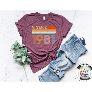May include: A maroon t-shirt with a vintage 1981 design featuring a sunset and rainbow stripes. The text "Vintage" is above the year.