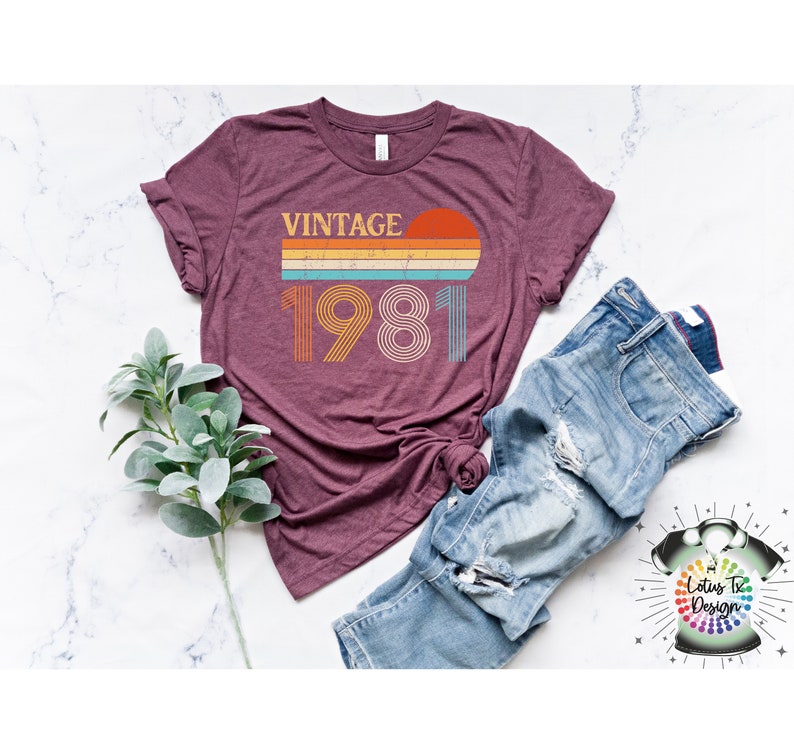 May include: A maroon t-shirt with a vintage 1981 design featuring a sunset and rainbow stripes. The text "Vintage" is above the year.