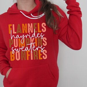 May include: A red hoodie with a white drawstring and a front pocket. The sweatshirt displays the words "FLANNELS, hayrides, PUMPKINS, sweaters, BONFIRES" in autumnal colours.