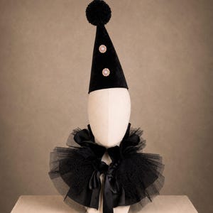 A mannequin head wearing a black party hat with two small gold-colored embellishments, with pom-poms on top, with the mannequin head wearing a black tulle collar, and a black bow tie.