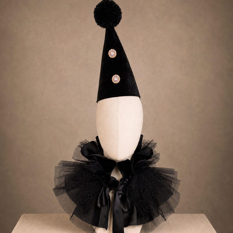 A mannequin head wearing a black party hat with two small gold-colored embellishments, with pom-poms on top, with the mannequin head wearing a black tulle collar, and a black bow tie.