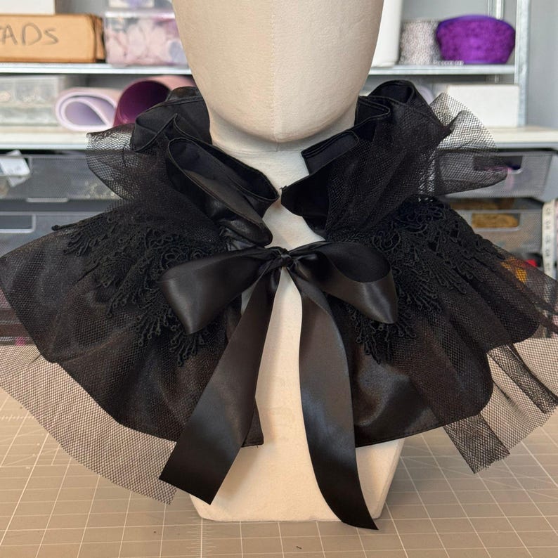 A mannequin head wearing a black lace and black satin cape with a black ribbon bow.