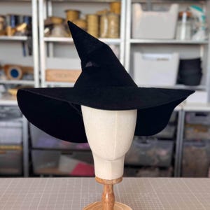 May include: A black witch hat with a tall, pointed crown and a wide, flexible brim. The hat is displayed on a beige mannequin head stand, showcasing the solid black fabric texture and the structured, slightly asymmetrical shape of the pointed top.